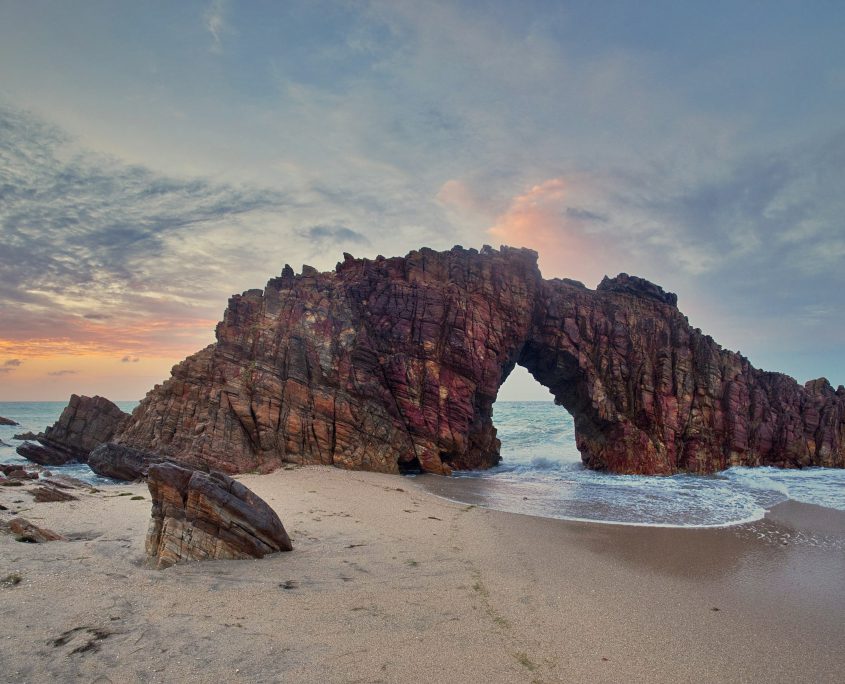 Pedra furada ao entardecer, o céu com núvens e alaranjado.